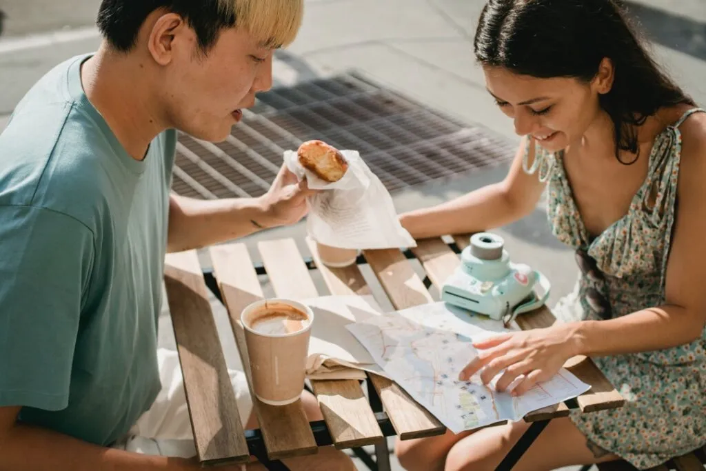 Two people sit at an outdoor table with coffee, a pastry, a map, and a mint-green instant camera.
