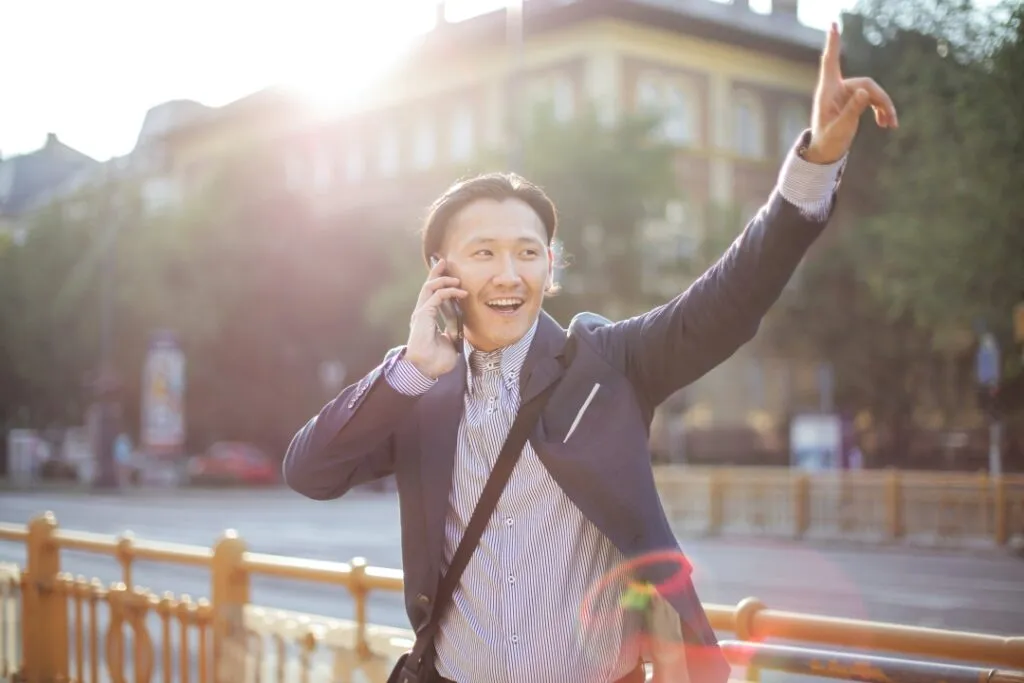 A man in a suit gestures while talking on a phone outdoors, with sunlight and urban surroundings.