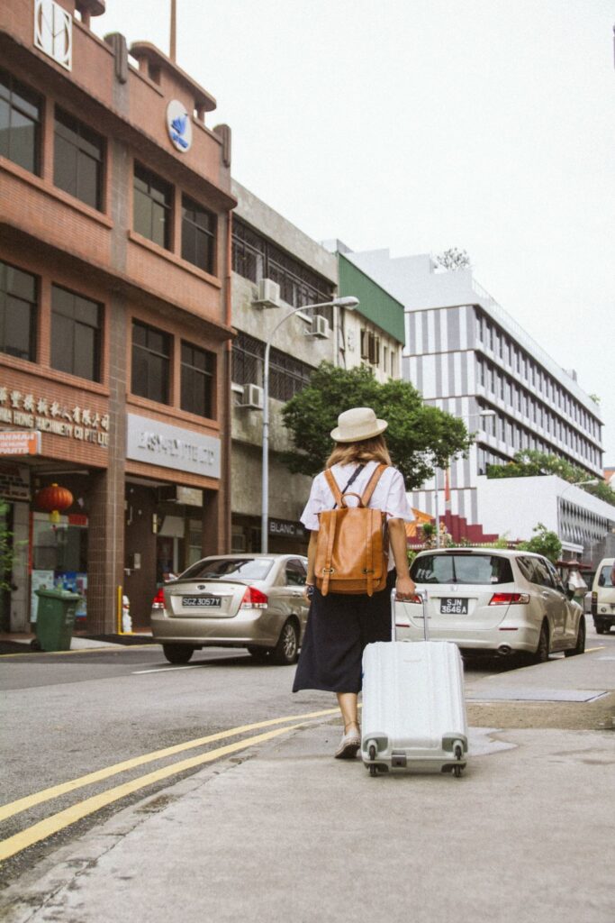 A traveler walks along a city street with a white suitcase and a brown backpack, surrounded by cars.