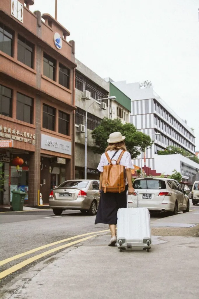 A traveler walks along a city street with a white suitcase and a brown backpack, surrounded by cars.