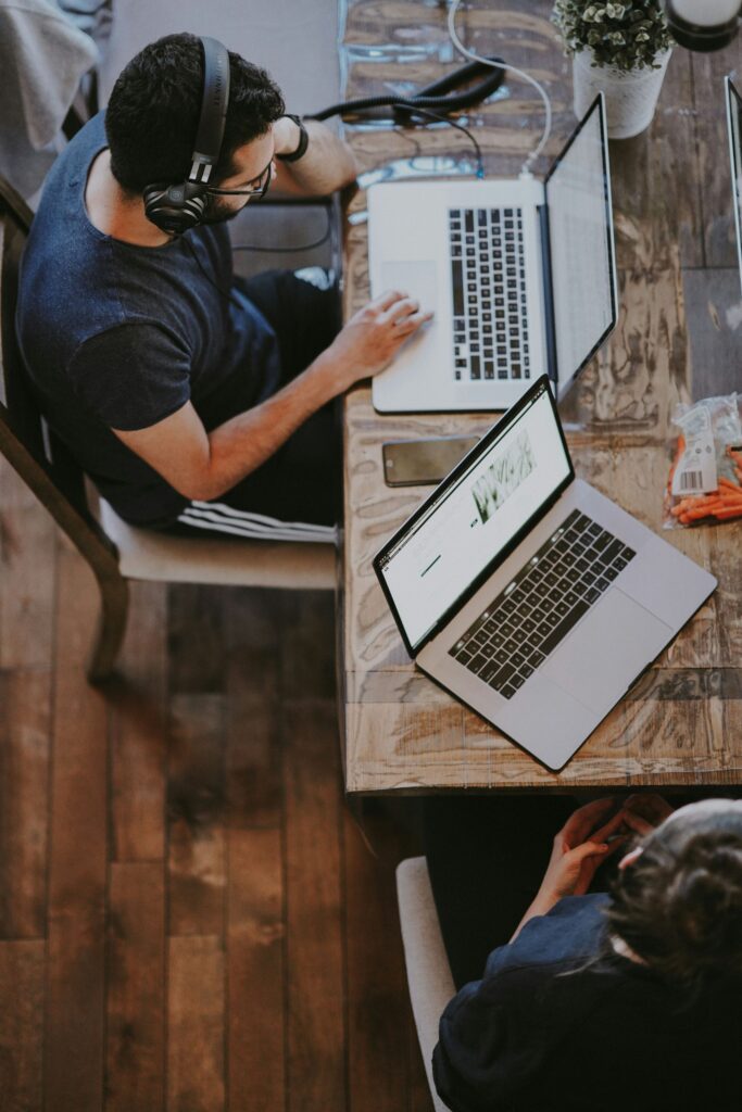 Two people work at a wooden table with laptops, headphones, and snacks in a casual setting.