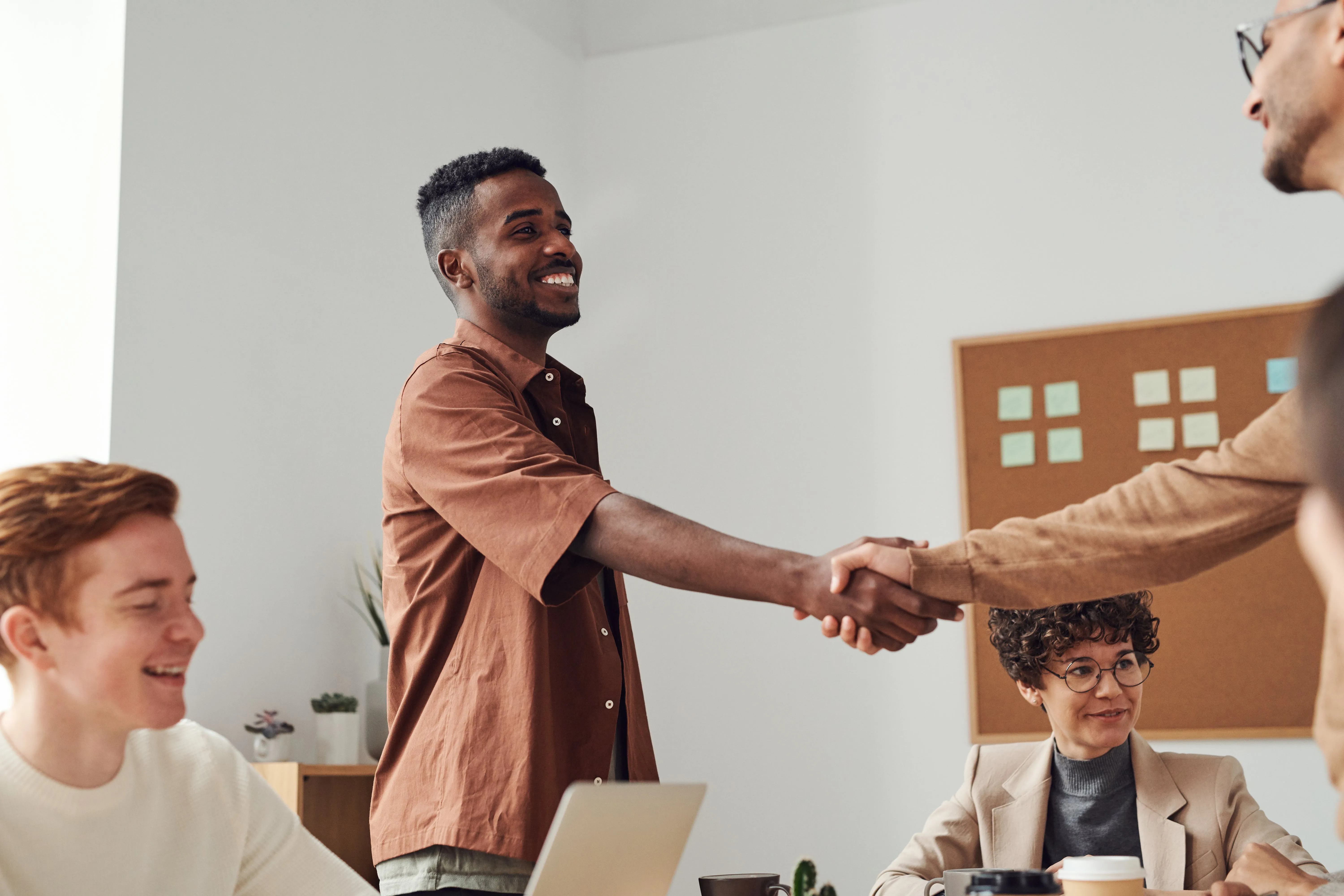 Two individuals are shaking hands in a professional meeting setting, surrounded by colleagues.