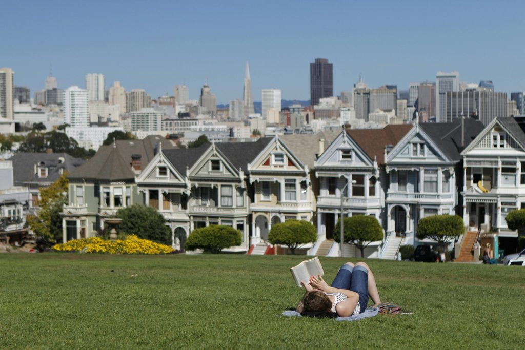 A person reads on grass near San Francisco's Painted Ladies, with the city skyline in the background.