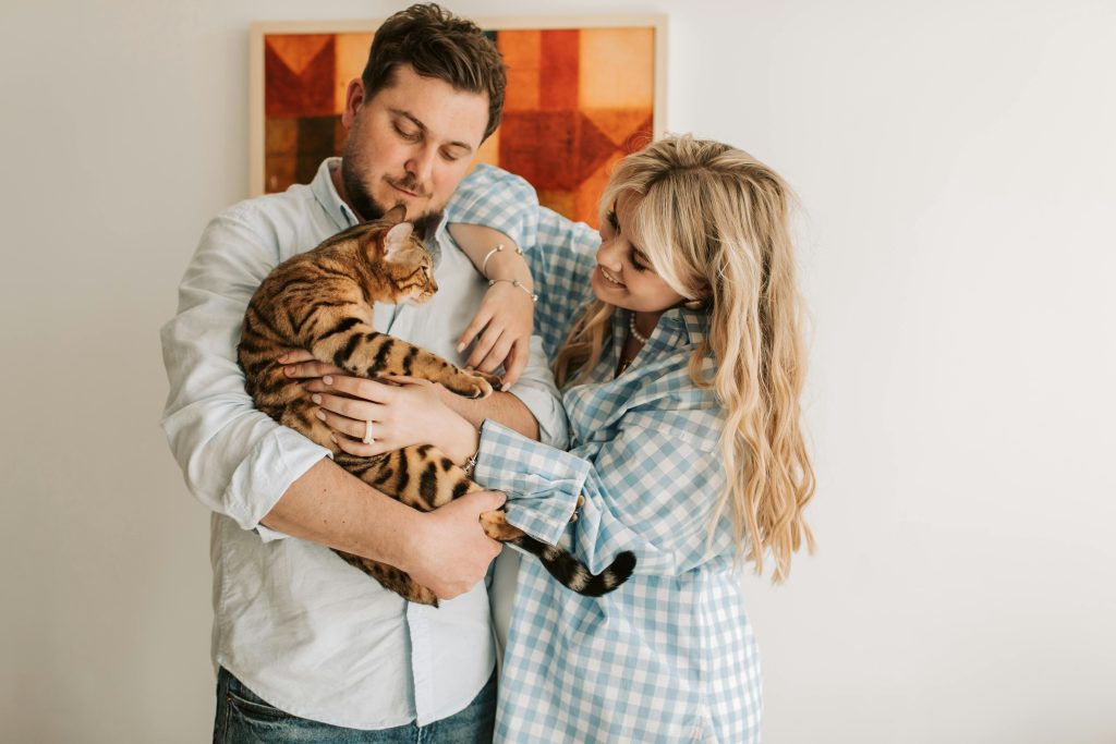 A couple holds a Bengal cat indoors, with a geometric artwork displayed on the wall behind them.