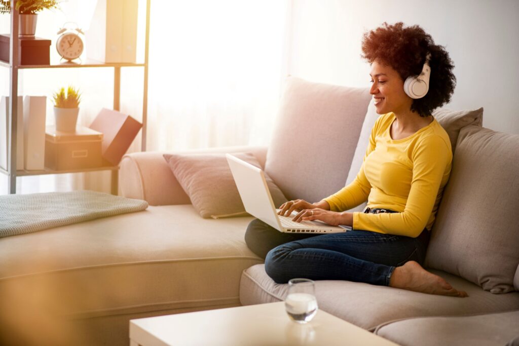 A person wearing headphones works on a laptop while seated on a cozy sofa in a sunlit room.