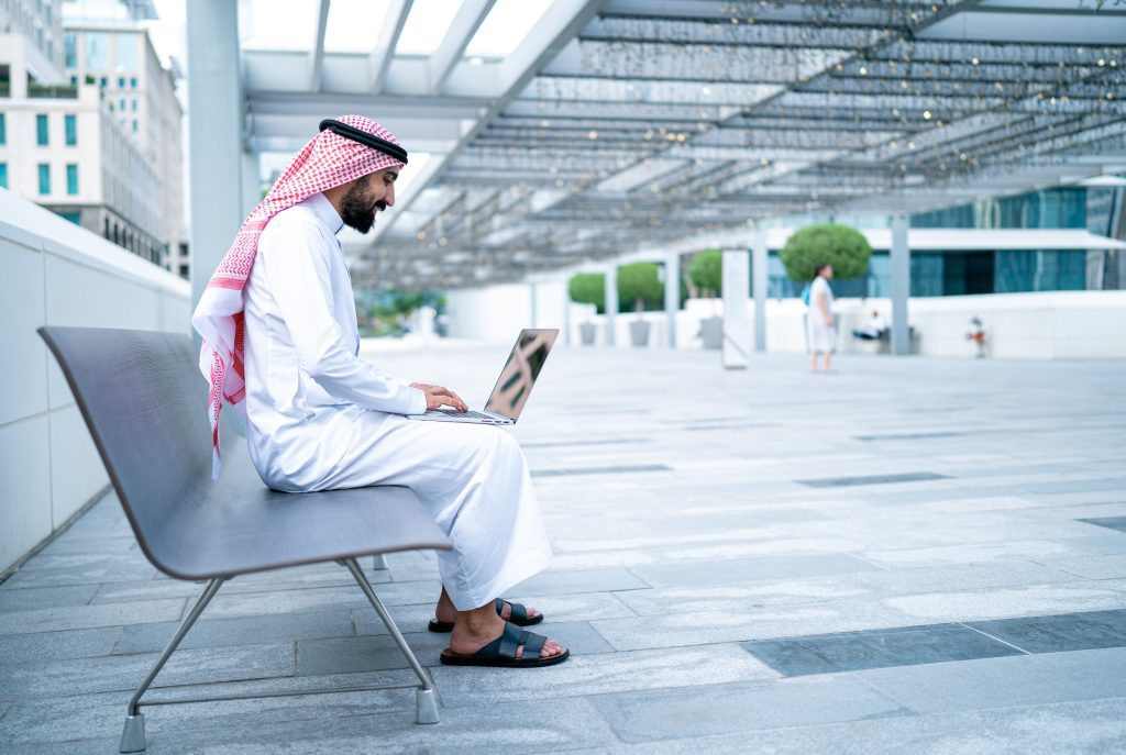 A man in traditional Middle Eastern attire works on a laptop while seated on a modern outdoor bench.