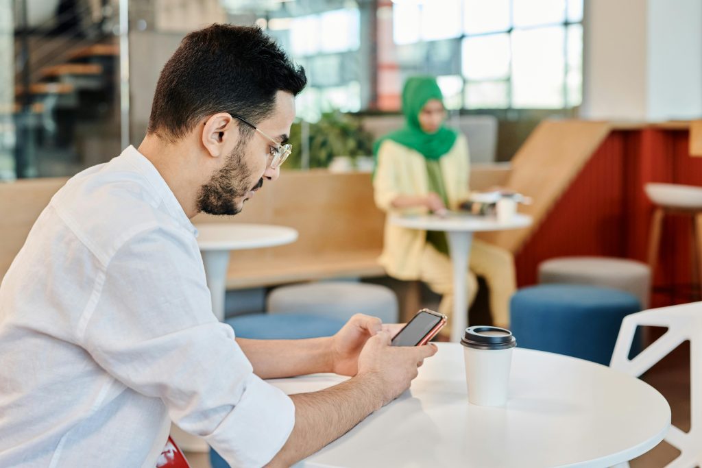 A man in a white shirt uses a smartphone at a cafe table with a coffee cup, while a woman sits nearby.