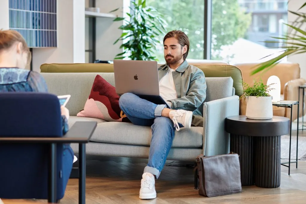 A person works on a laptop in a modern, well-lit lounge with plants and contemporary furniture.