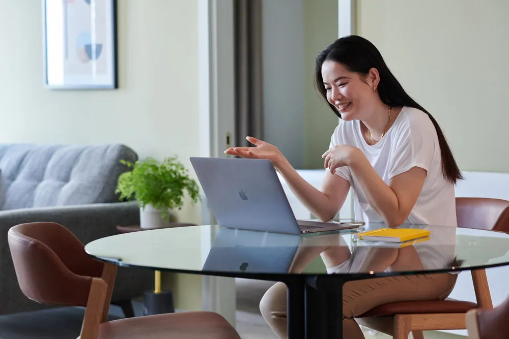 A woman sits at a glass table with a laptop, gesturing while engaged in a conversation or meeting.