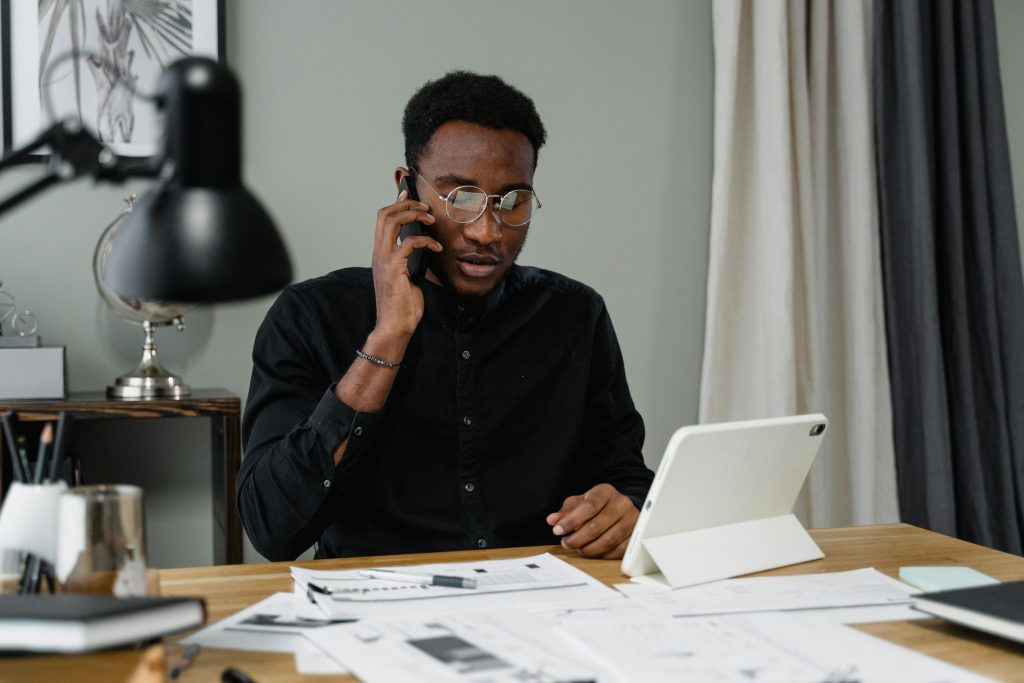 A person in a black shirt sits at a desk with papers, a tablet, and a lamp, talking on the phone.