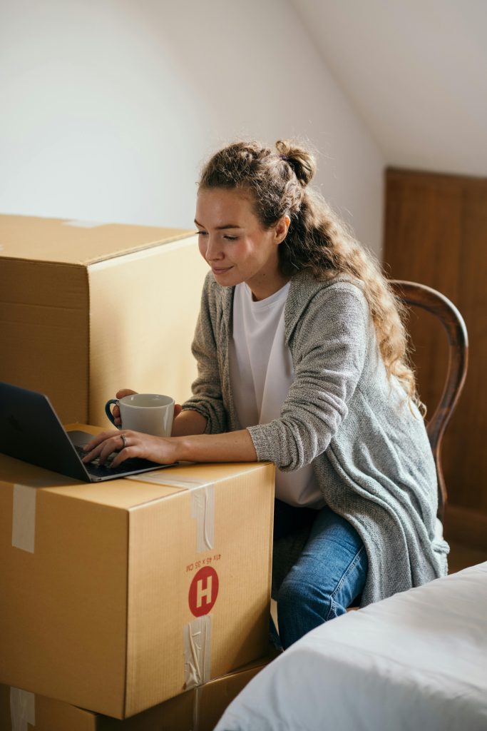 A woman works on a laptop while holding a mug, seated near cardboard boxes in a home setting.