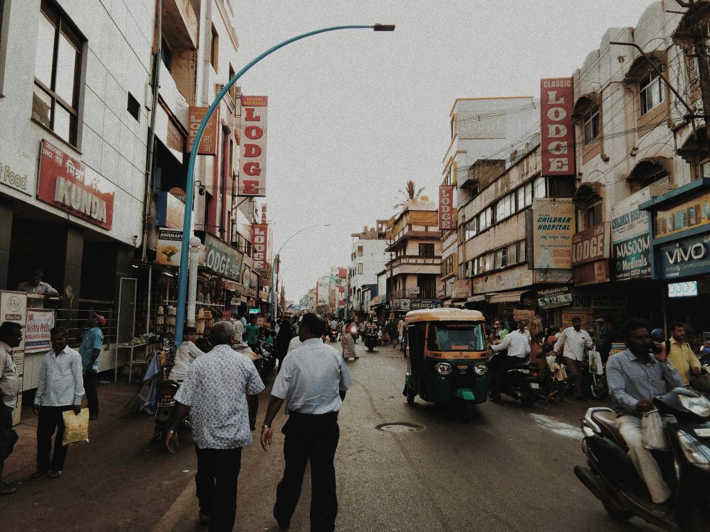 A bustling urban street with lodges, shops, pedestrians, and vehicles, including an auto-rickshaw.