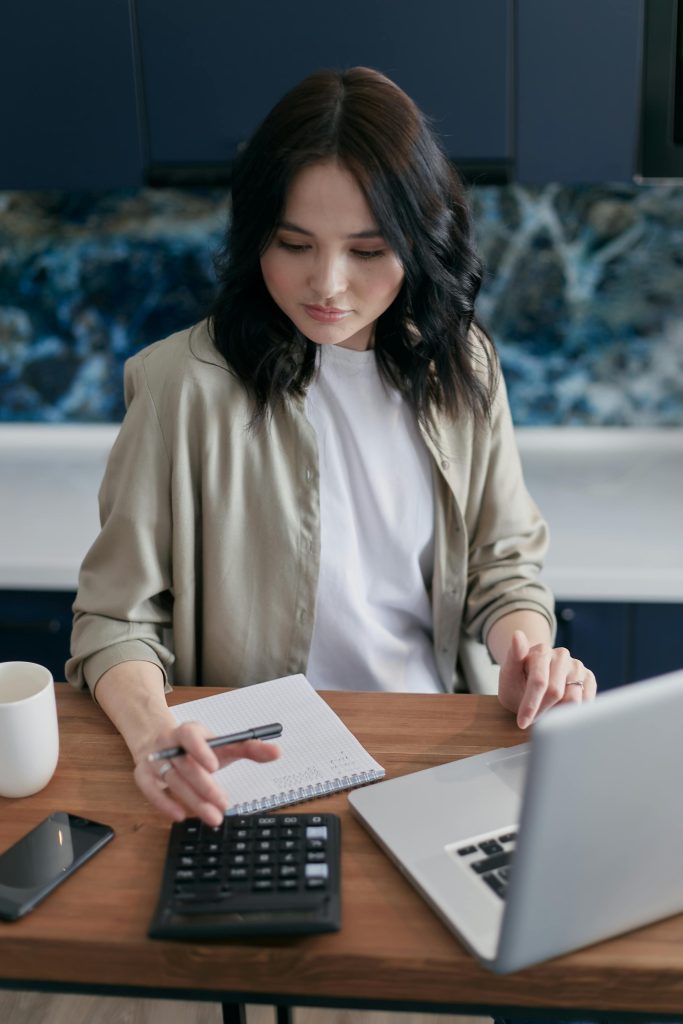 A person works at a desk with a calculator, notebook, laptop, smartphone, and coffee mug.