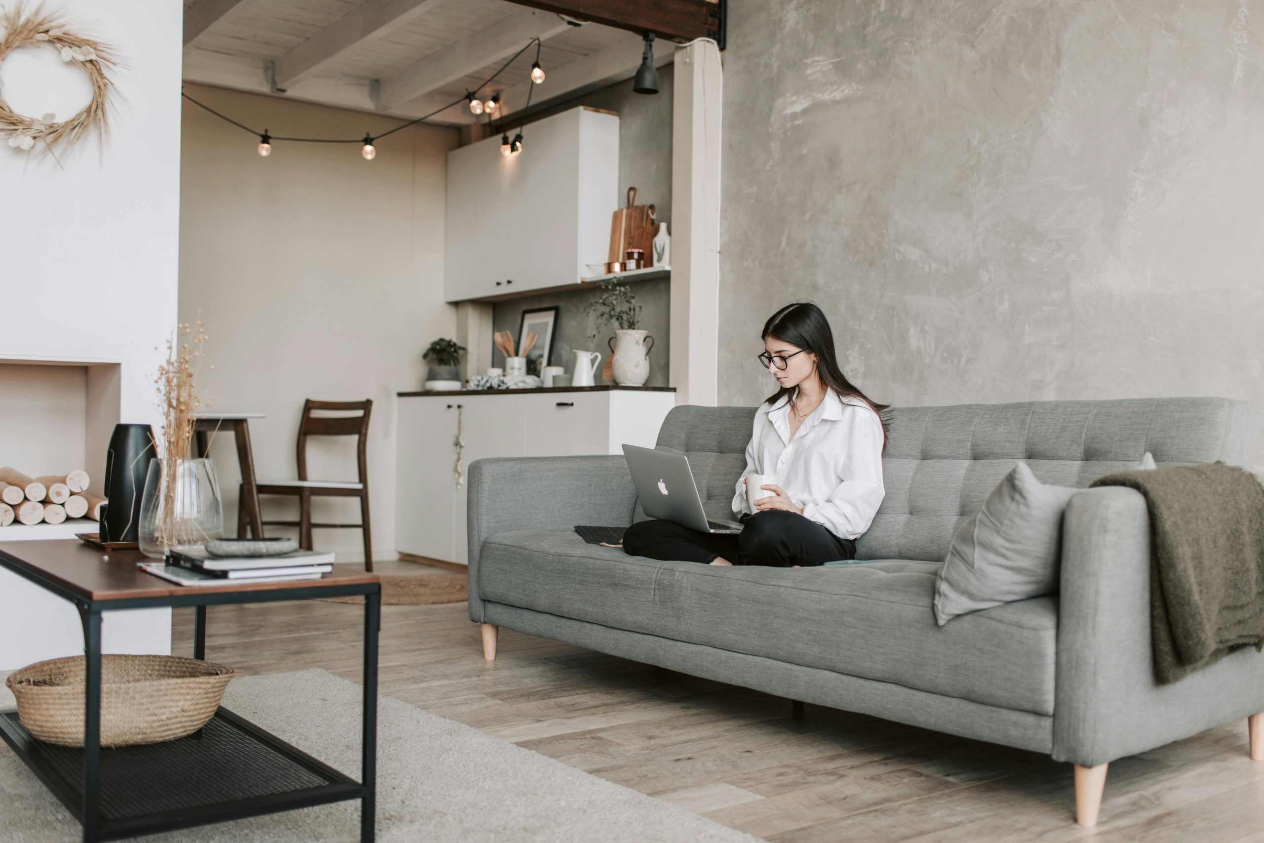 A modern living space with neutral tones features a woman seated on a gray sofa using a laptop.