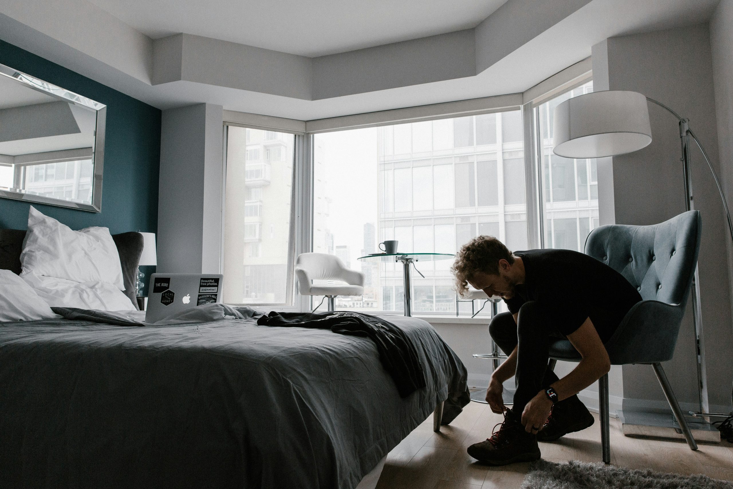 A modern bedroom with large windows, a person tying shoes, and a laptop on the bed.
