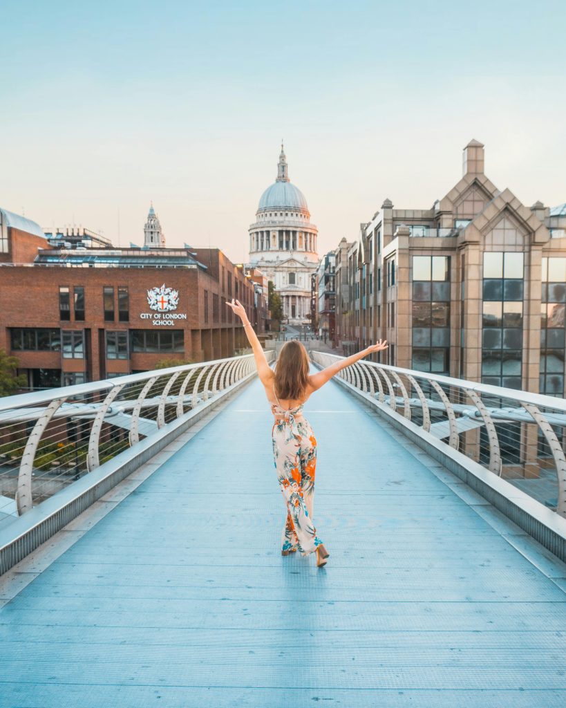 A woman in a floral dress walks joyfully on Millennium Bridge, with St. Paul's Cathedral in view.