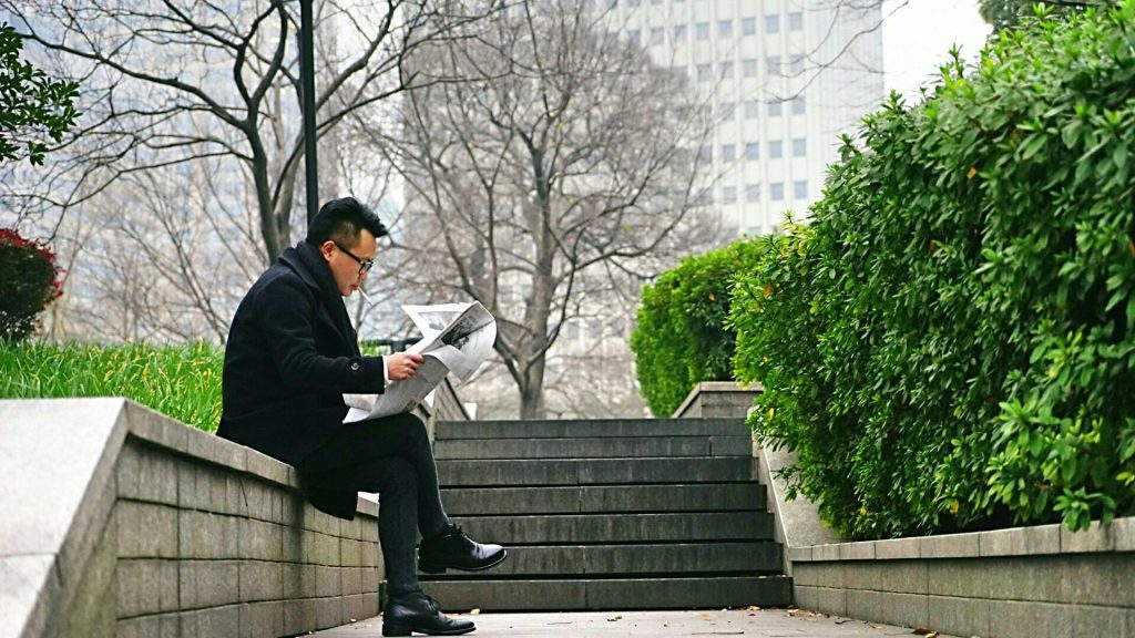 A man in dark attire reads a newspaper while seated on outdoor steps surrounded by greenery.