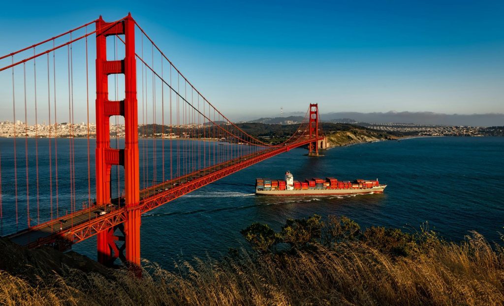 Golden Gate Bridge spans blue waters as a cargo ship passes beneath, framed by hills and cityscape.
