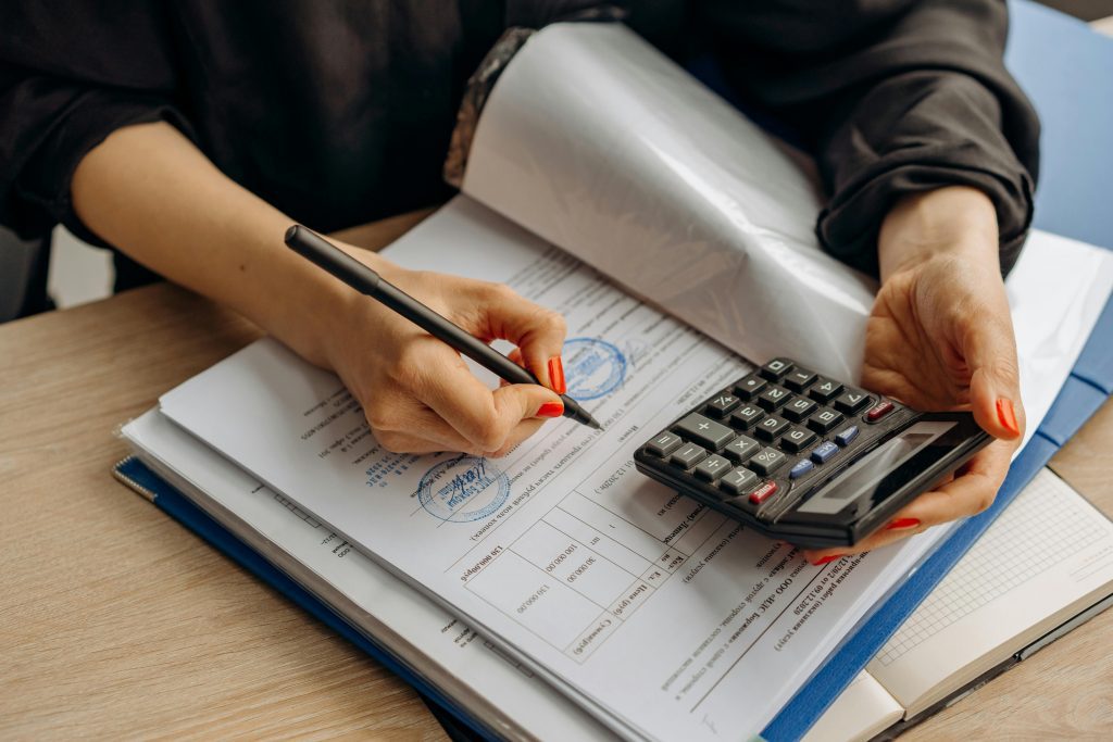 A person calculates and signs documents at a desk, holding a pen and a calculator.