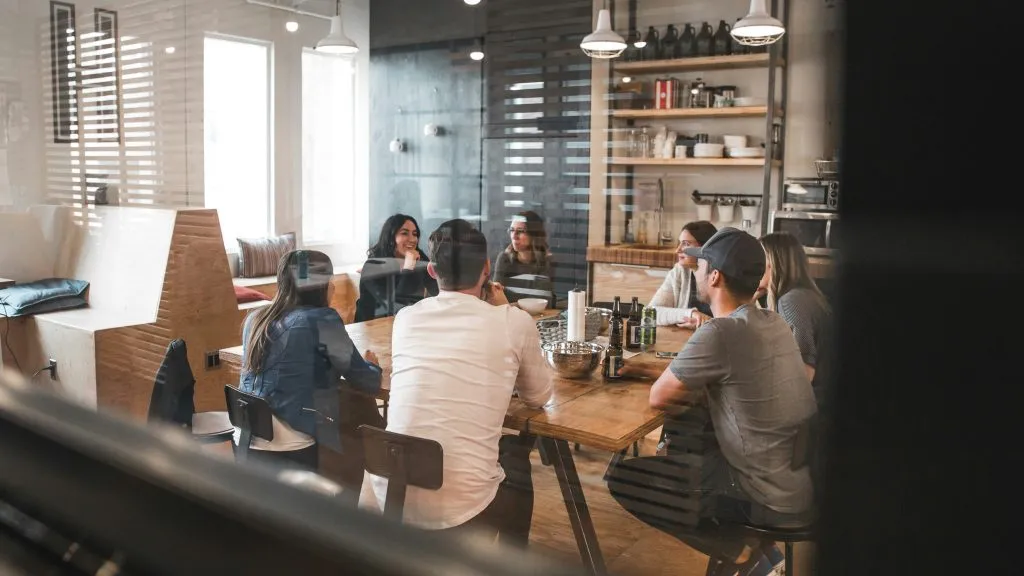 A group of people sits around a wooden table in a modern, well-lit room with shelves and decor.