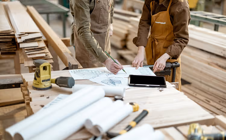 2 people planning blueprints in a large workshop with wooden planks lying around.