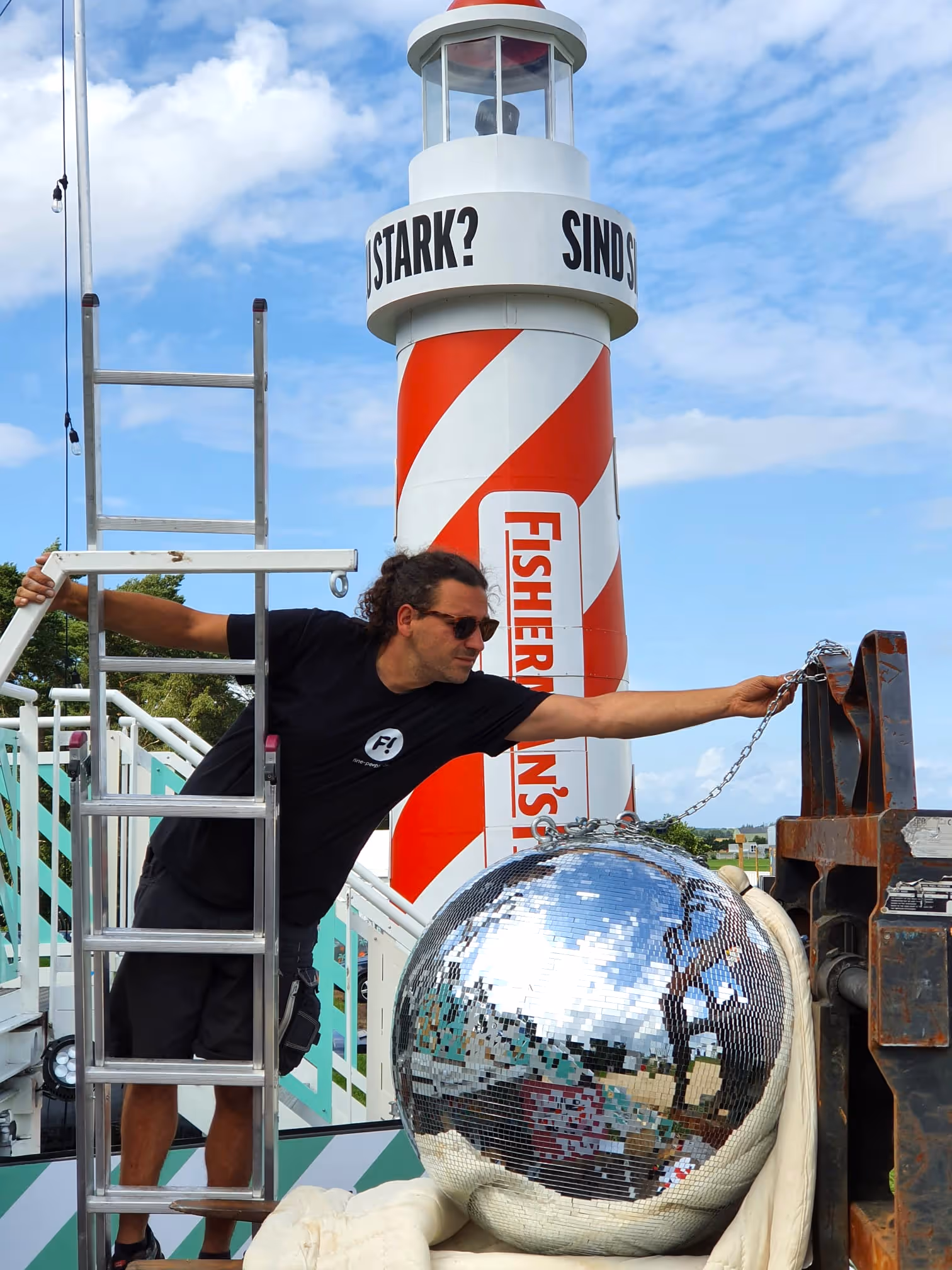 Mann mit Sonnenbrille lehnt sich an eine Leiter und hält eine Kette, die an einer großen Discokugel hängt, mit einem rot-weißen Leuchtturm im Hintergrund.