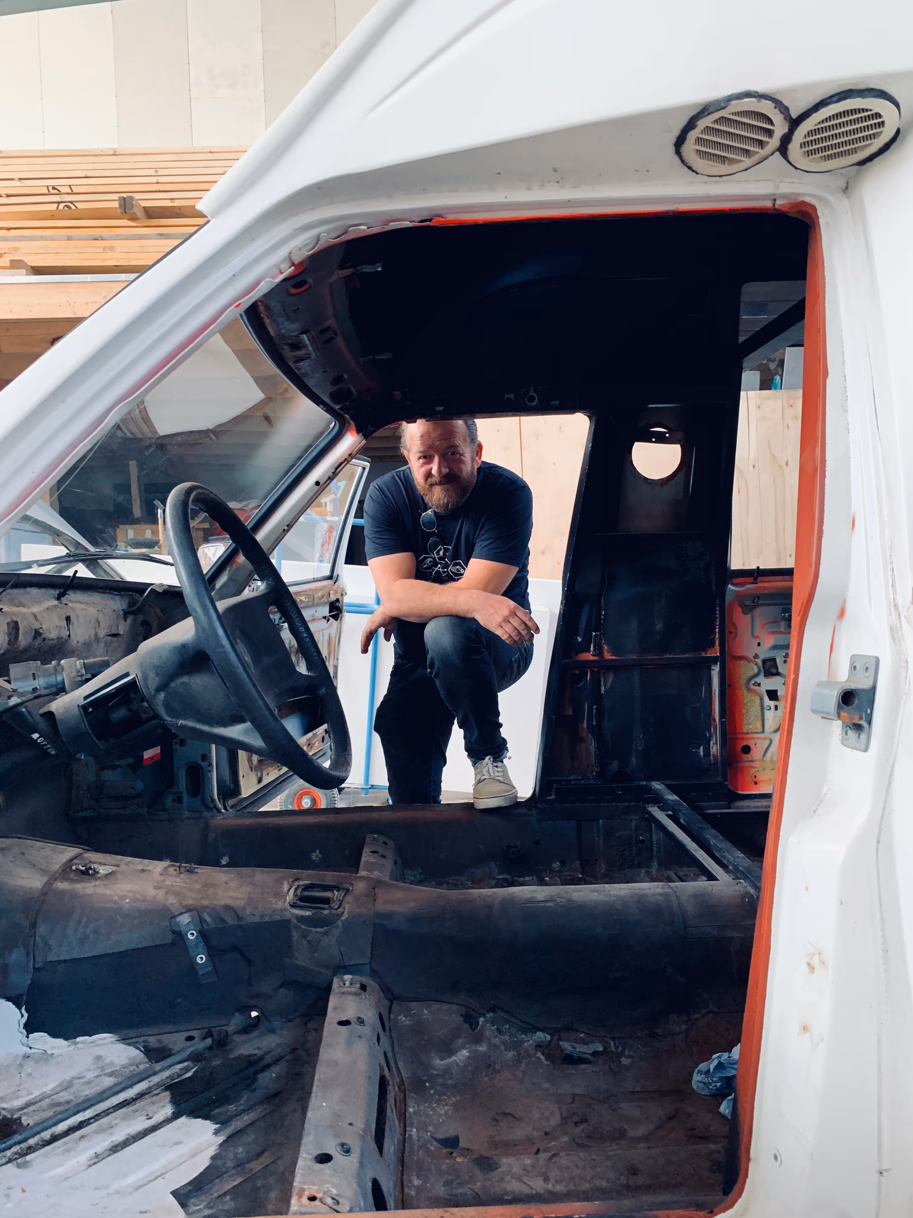 Mann mit Bart kniet neben dem leergeräumten Innenraum eines alten Autos ohne Sitze und Armaturenbrett.