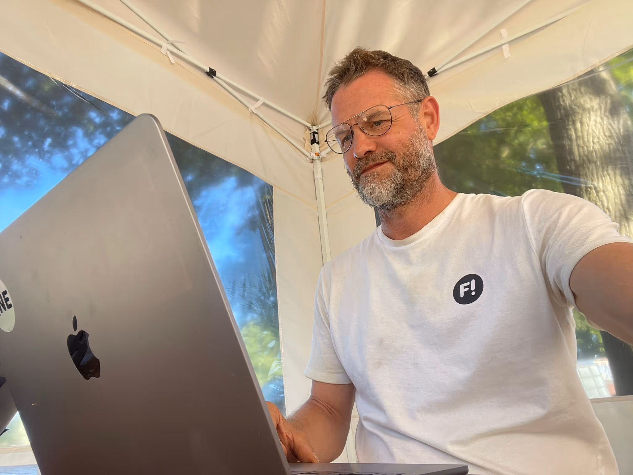 Mann mit Brille und Bart arbeitet an einem Laptop unter einem weißen Zelt mit Bäumen im Hintergrund.
