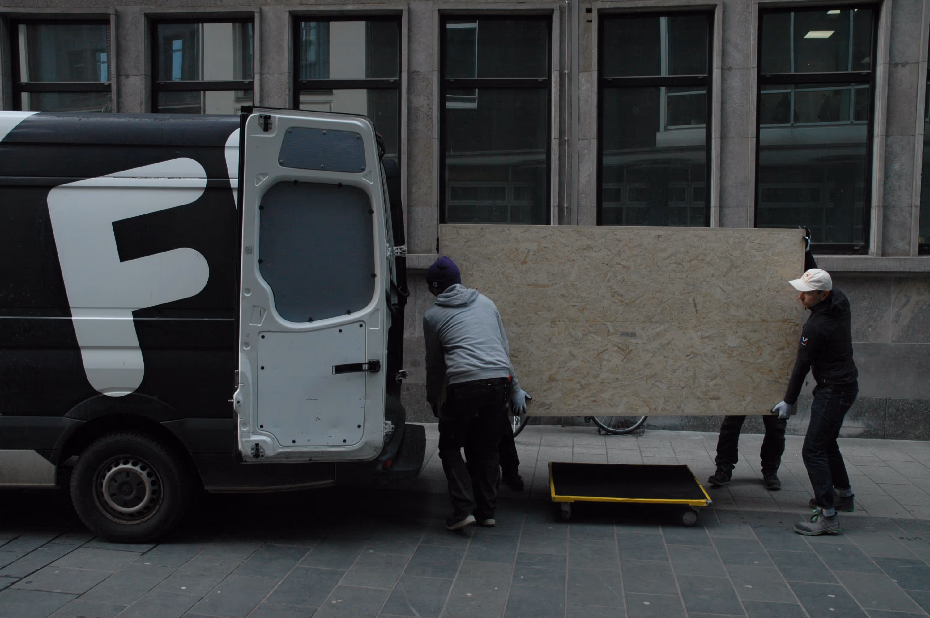 Drei Männer tragen ein großes Holzbrett aus einem Lieferwagen auf einen Rollwagen auf einem städtischen Bürgersteig.