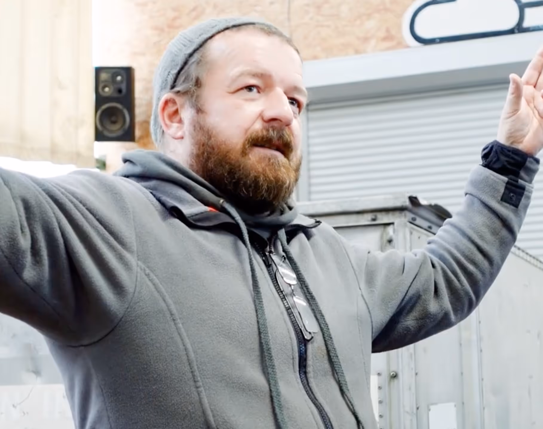 Mann mit grauer Strickmütze und Bart, der in einem grauen Kapuzenpullover die Arme hebt.