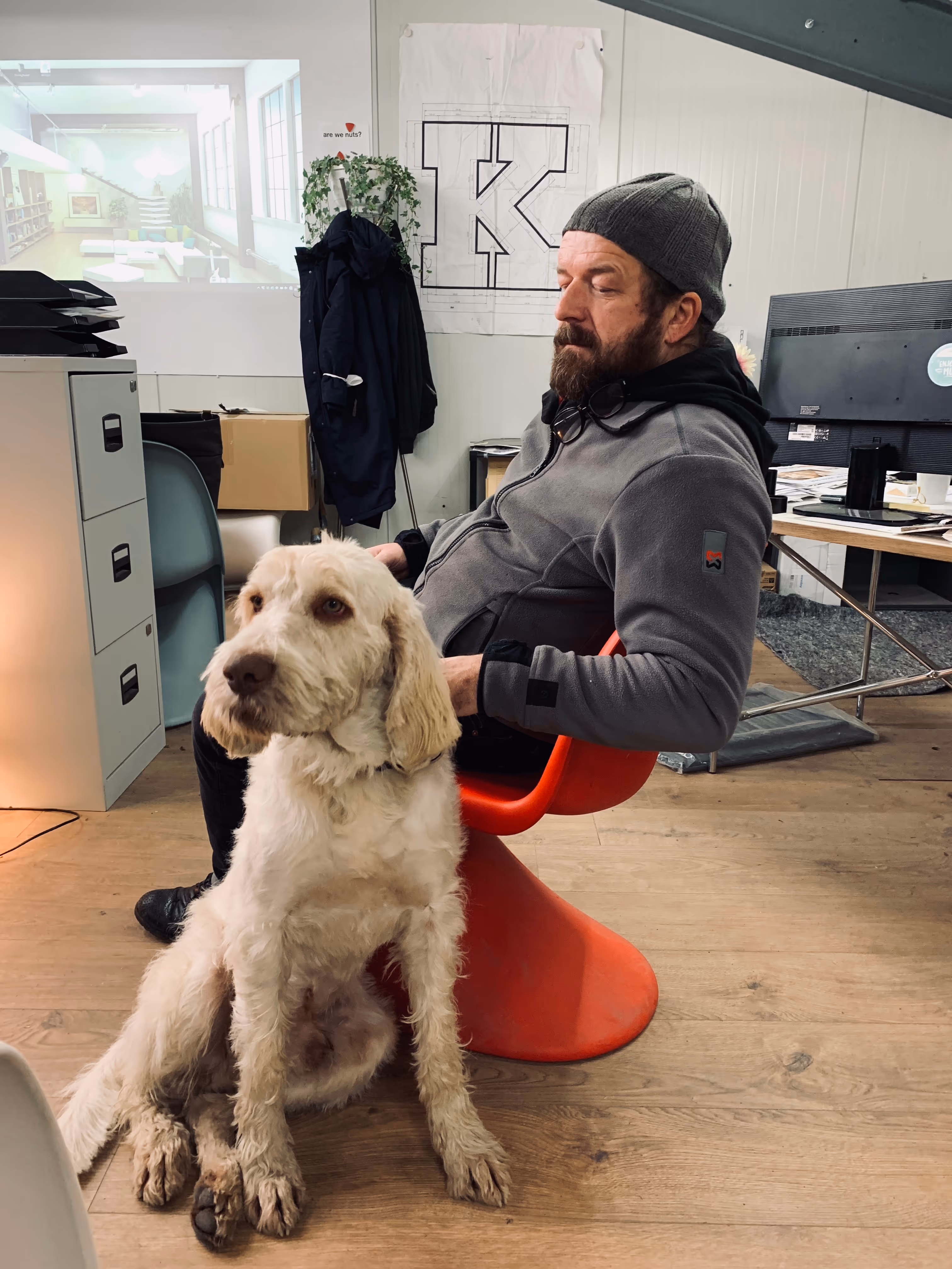 Mann mit grauer Mütze und Kapuzenpullover sitzt auf rotem Stuhl im Büro, vor ihm sitzt ein großer weißer Hund.