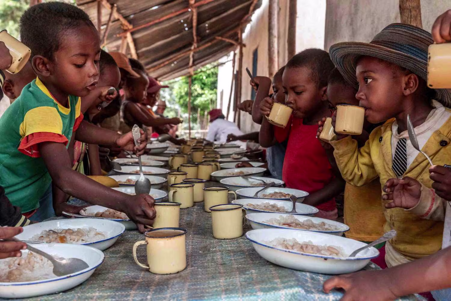 Children seated on both sides of a long table drinking from yellow mugs and eating food from white enamel bowls.