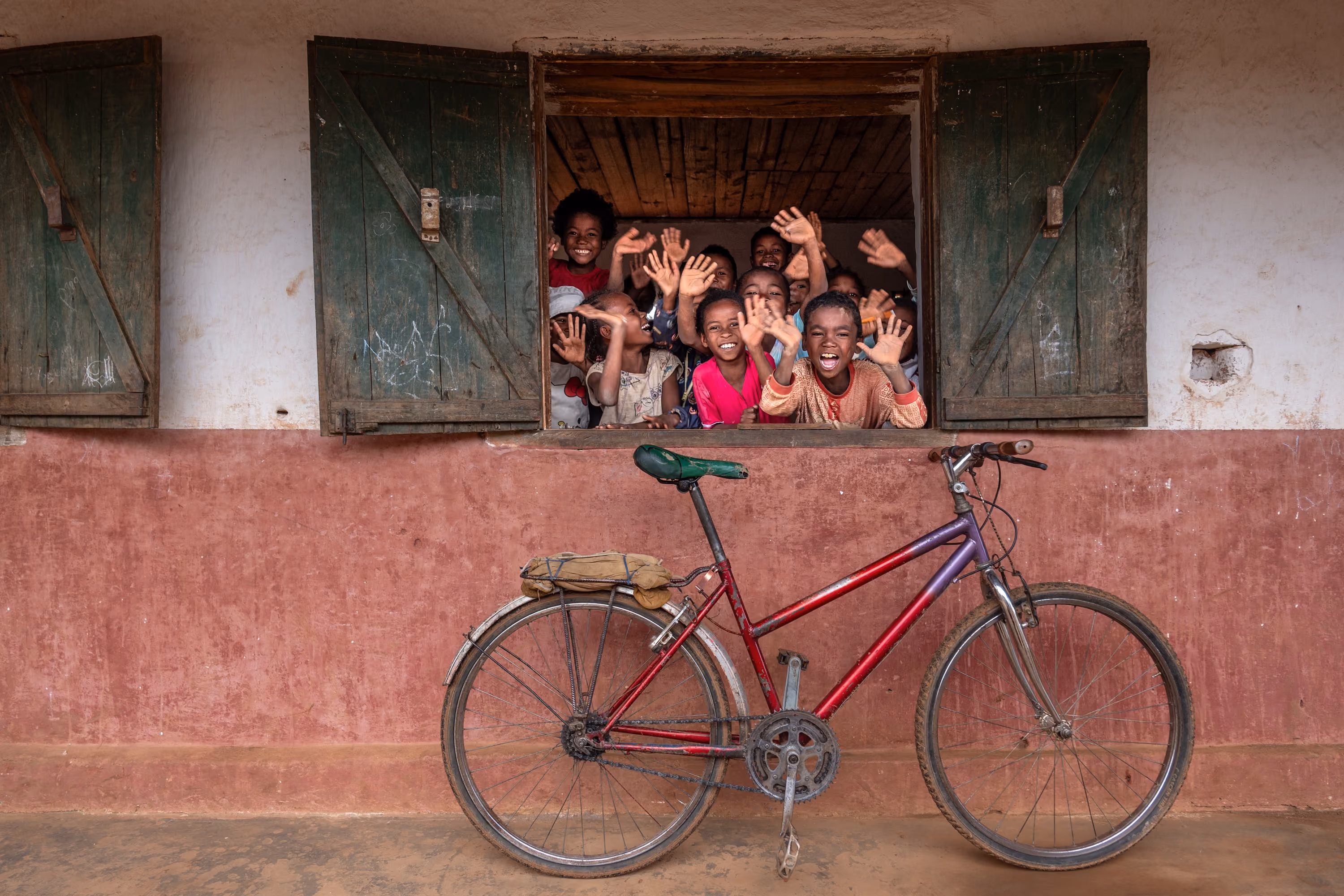 A bike rests against a brick wall under the window where kids are waiving to the camera.
