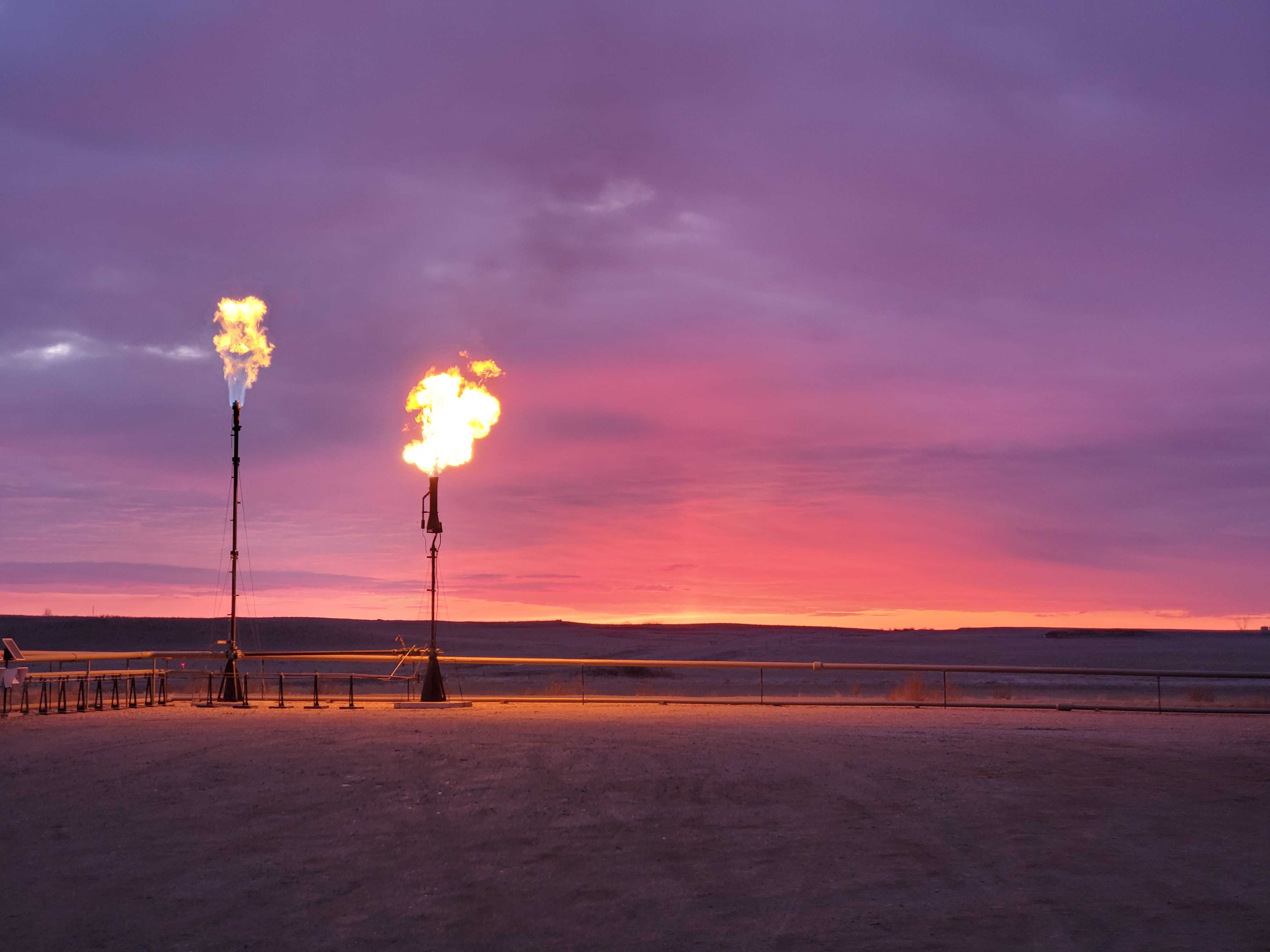Two oil-field flare stacks burning excess methane against a purple sunset sky, illustrating Crusoe’s flare gas capture for powering data centers.