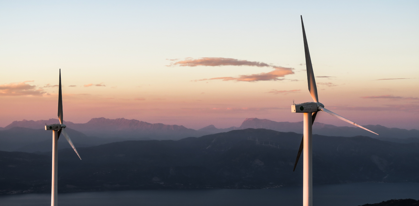 Two wind turbines at sunset overlooking mountains and a lake, symbolizing Crusoe’s use of renewable energy.