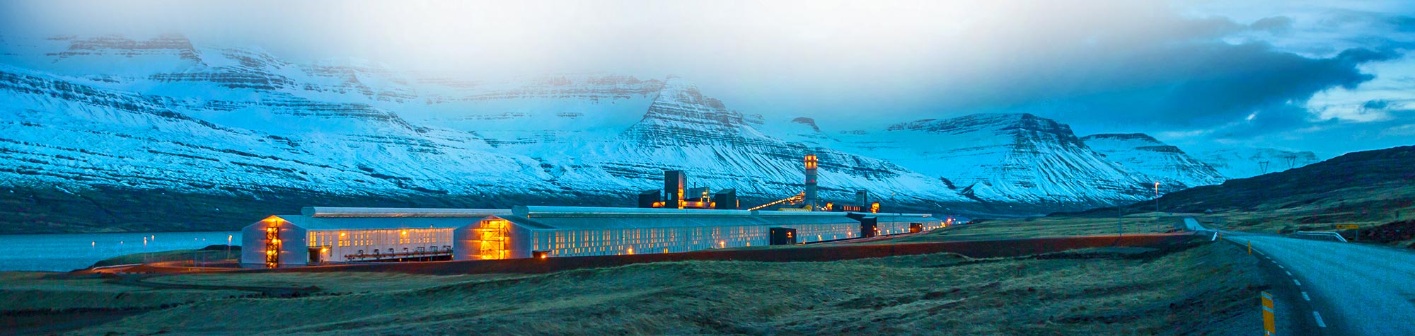 A large industrial facility illuminated with warm lights against snowy mountains in the background.