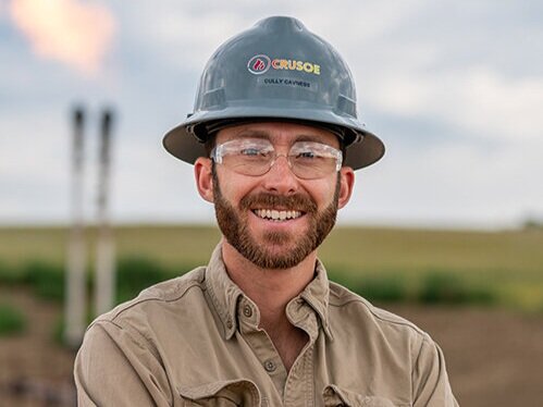 An image of Cully Cavness, Co-founder and COO of Crusoe, smiling while wearing a company-branded hard hat and safety glasses.