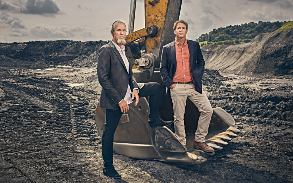 Two individuals stand confidently next to heavy machinery at a mining site.