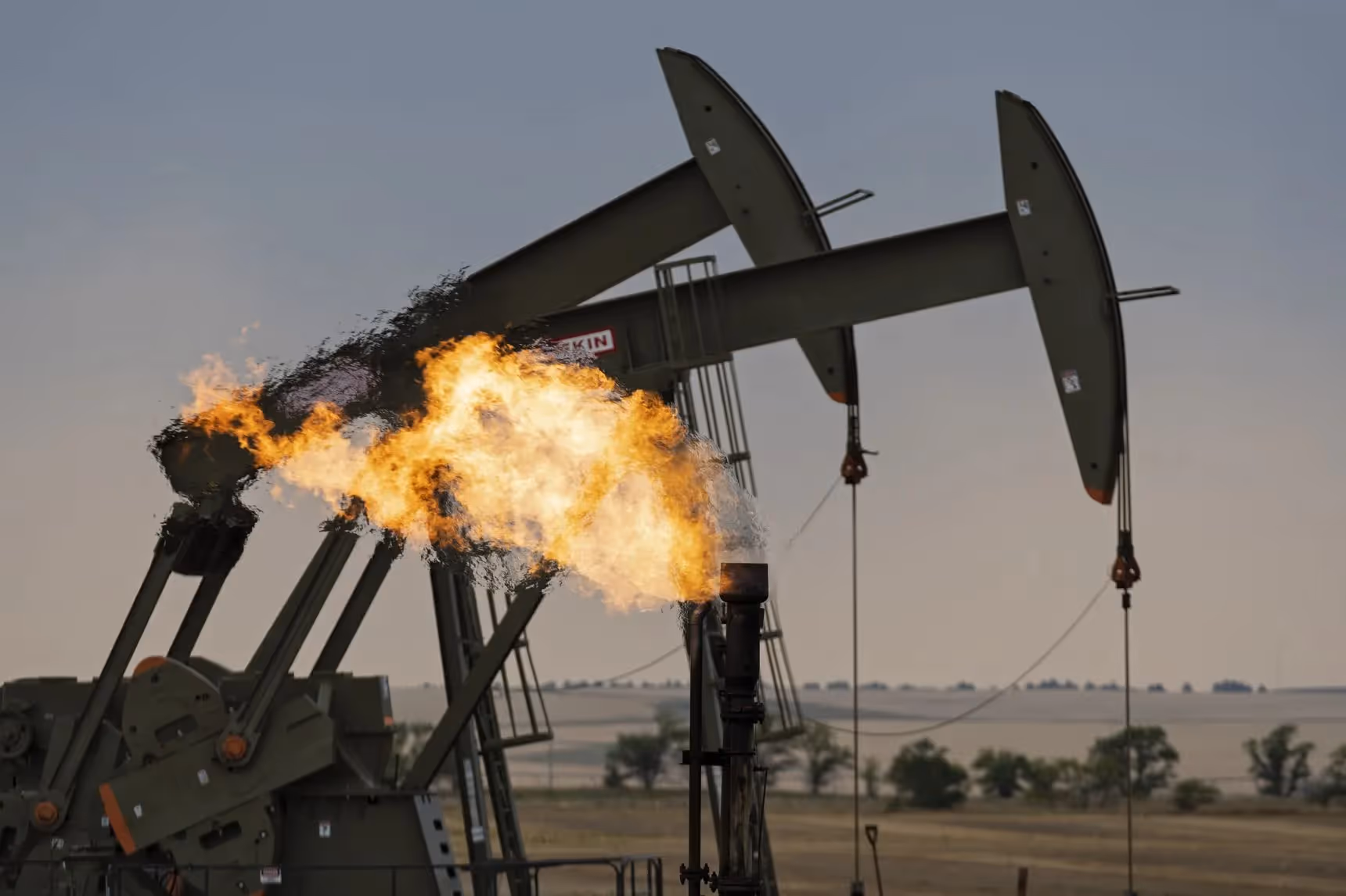 An oil pump jack operating in a rural landscape with a flare stack burning excess natural gas.