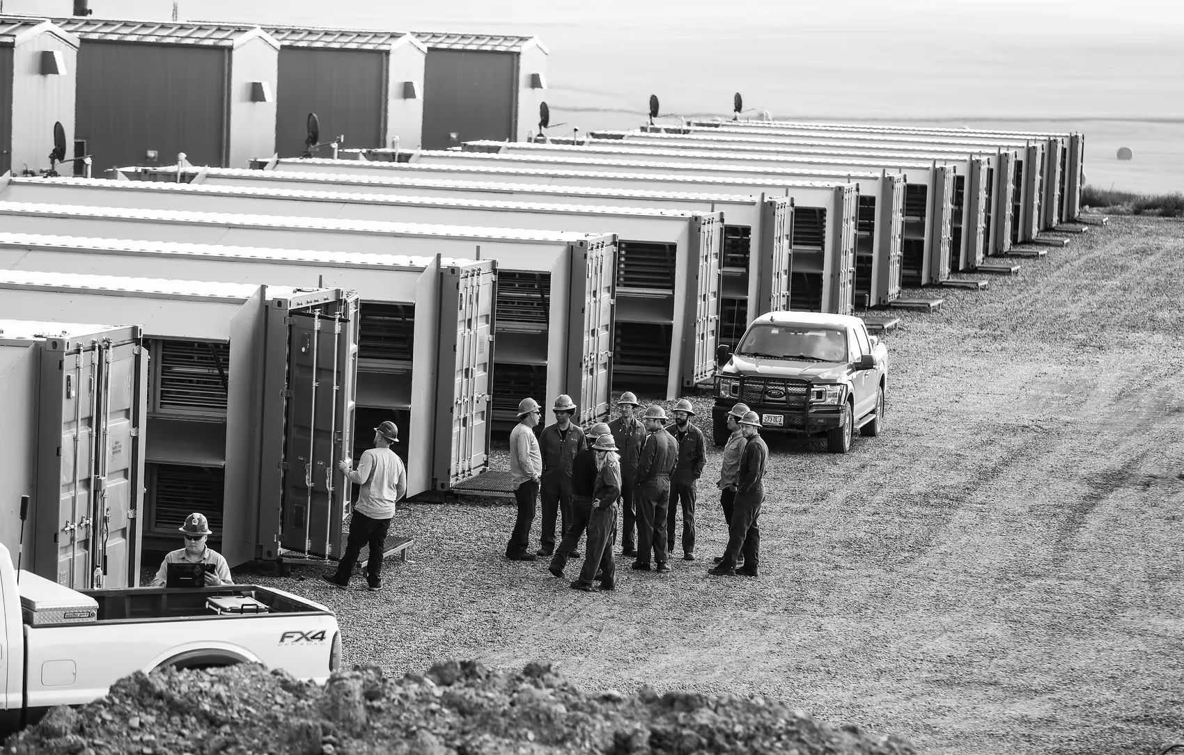 A black and white photograph of an industrial site where a team of workers in hard hats are gathered near rows of data.