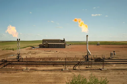 Wide-angle view of an oil and gas facility featuring two flare stacks emitting flames against a clear blue sky.
