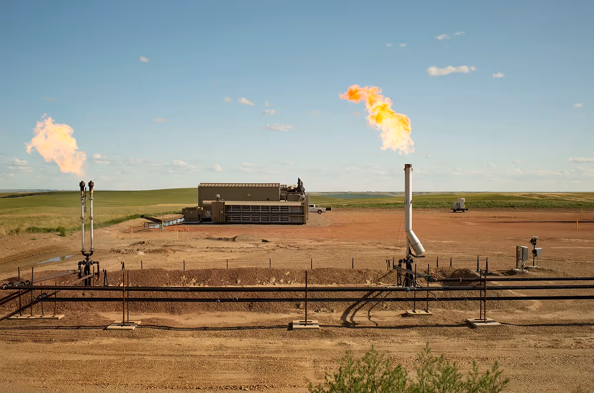 Wide-angle view of an oil and gas facility featuring two flare stacks emitting flames against a clear blue sky.