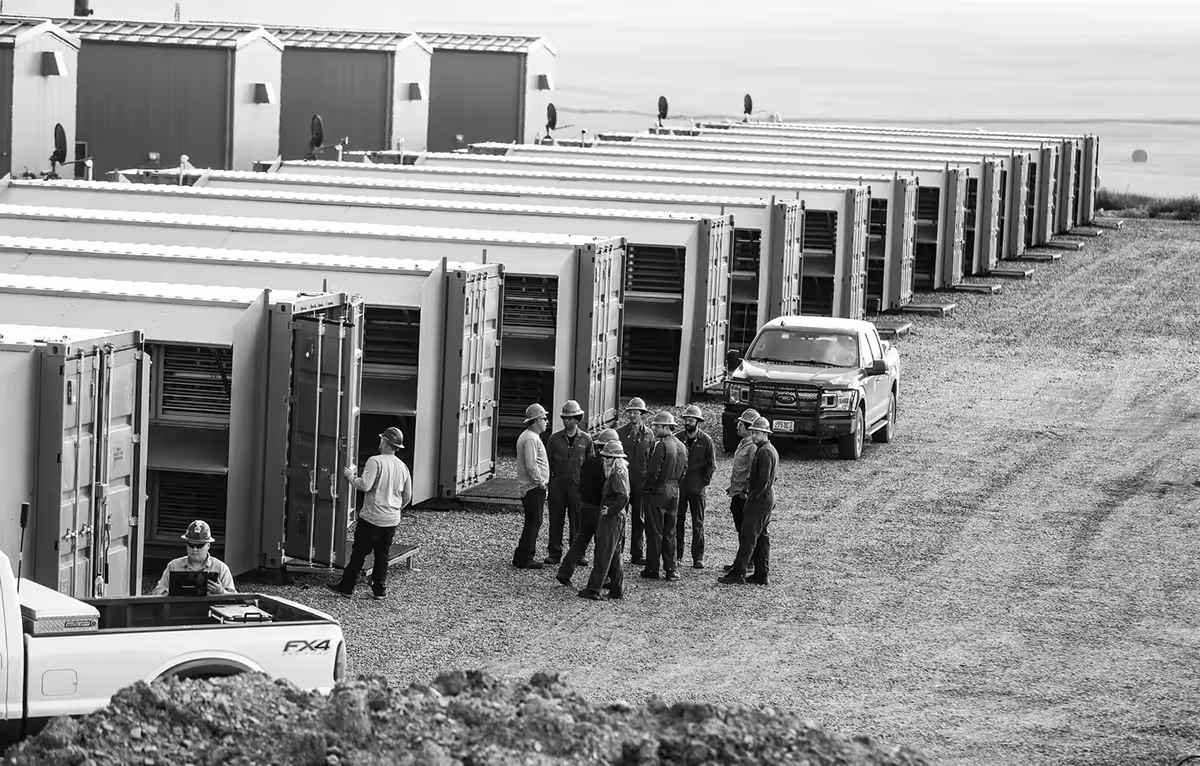 A black and white photograph of an industrial site where a team of workers in hard hats are gathered near rows of data.