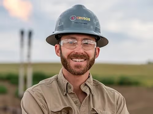 An image of Cully Cavness, Co-founder and COO of Crusoe, smiling while wearing a company-branded hard hat and safety glasses.