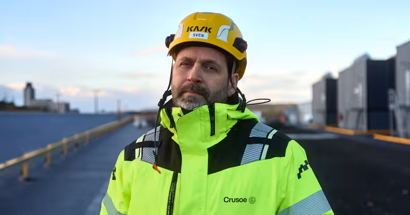 A construction worker wearing a yellow KASK helmet and Crusoe-branded high-visibility jacket stands at an outdoor job site.