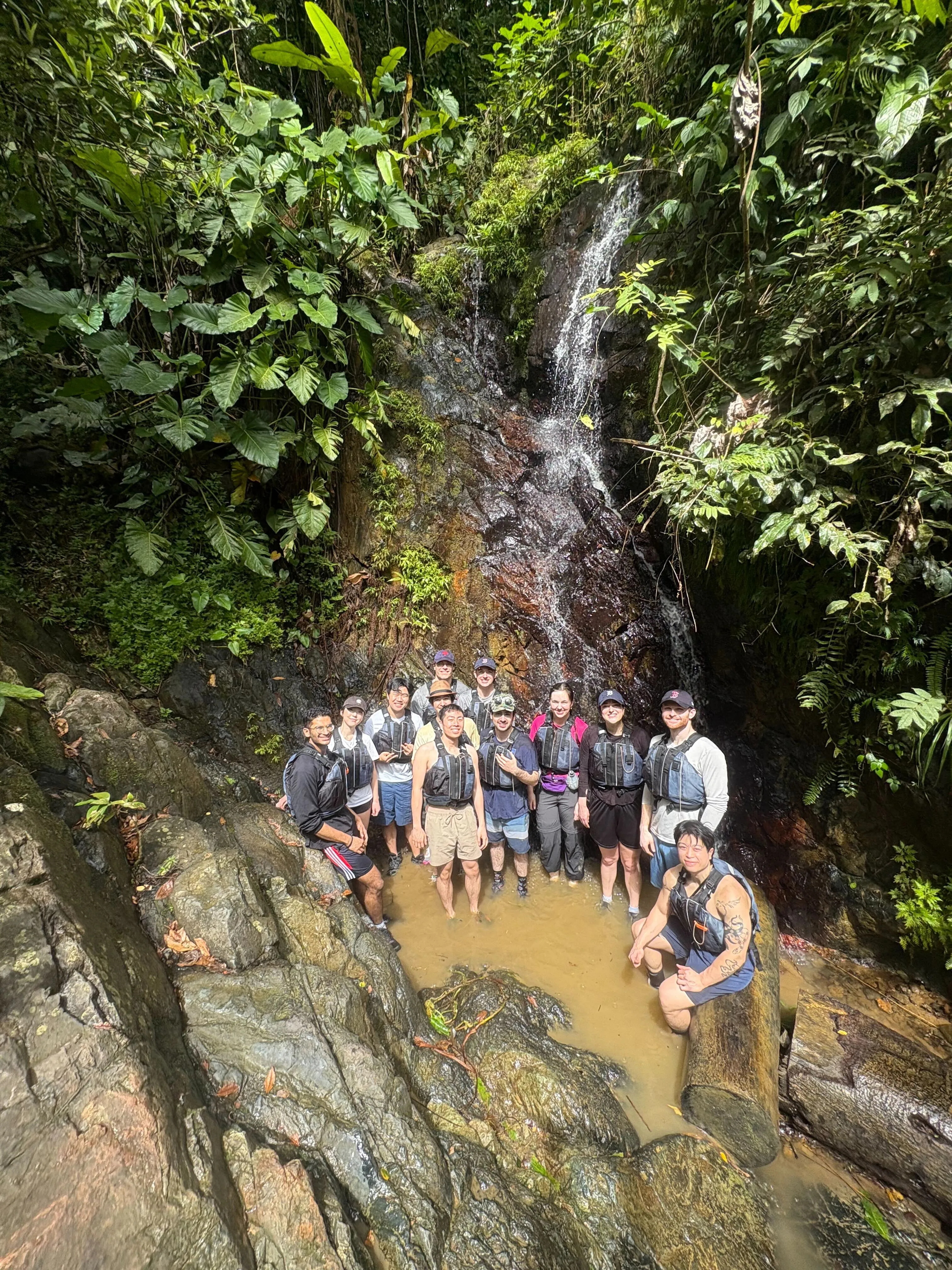 Group of people wearing life jackets standing in shallow water by a small waterfall in lush forest.