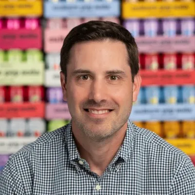 Smiling man with short dark hair and checked shirt in front of colorful blurred boxes.
