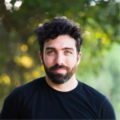 Man with dark curly hair and beard wearing a black shirt with green foliage background.