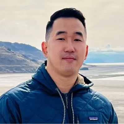 Man wearing a blue quilted jacket standing outdoors with a mountainous lake landscape behind him.