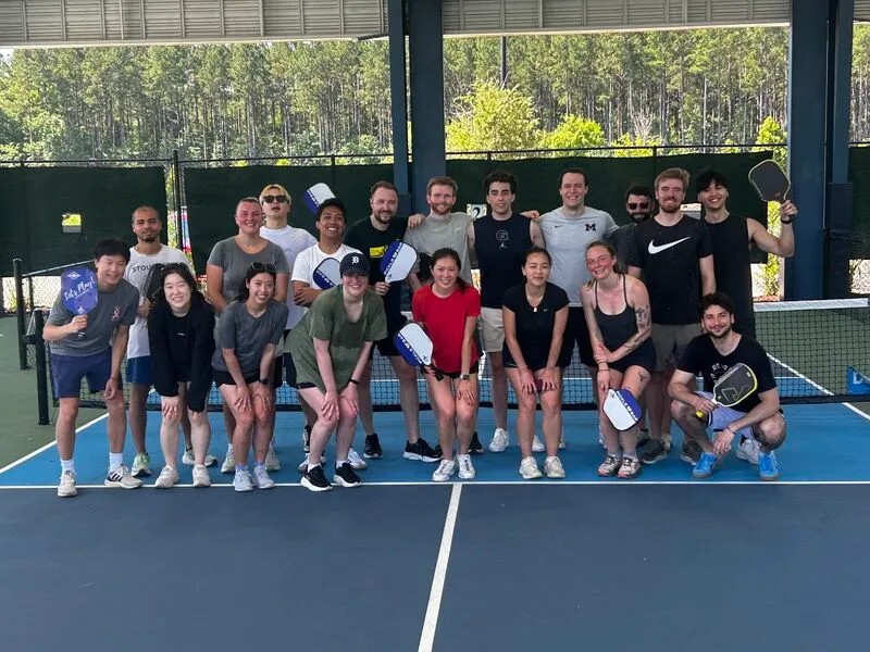 Group of 18 young adults posing on an outdoor pickleball court, holding paddles and smiling.
